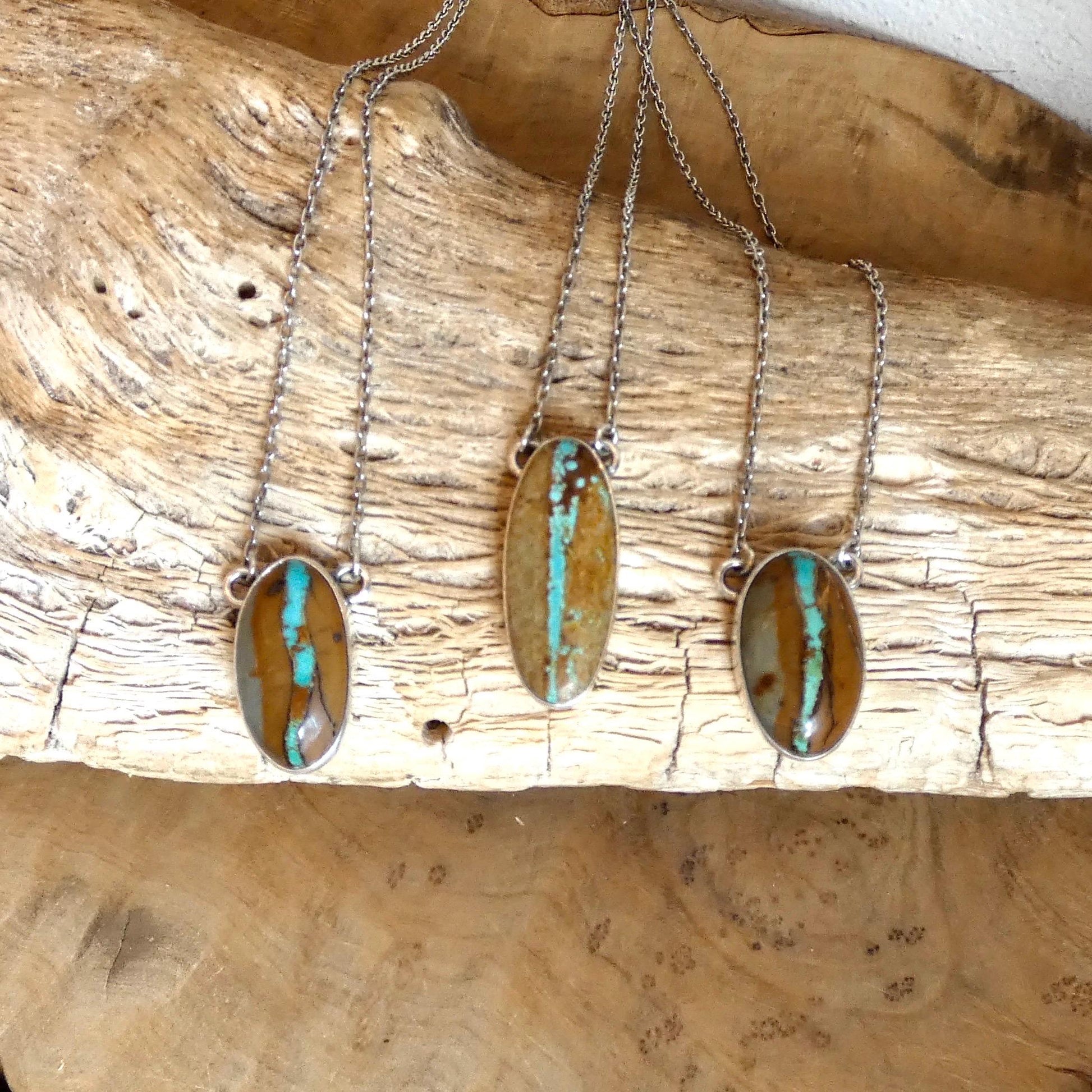 May include: Three silver necklaces with oval pendants. Each pendant features a brown and turquoise stone. The stones have a unique pattern of brown and blue-green streaks. The necklaces are displayed on a piece of wood.