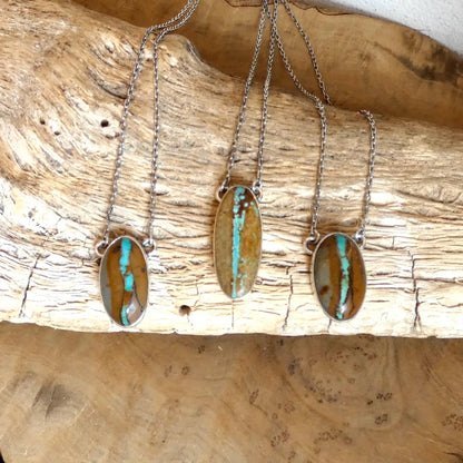 May include: Three silver necklaces with oval pendants. Each pendant features a brown and turquoise stone. The stones have a unique pattern of brown and blue-green streaks. The necklaces are displayed on a piece of wood.