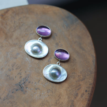 May include: A pair of silver earrings with a purple gemstone and a white iridescent shell. The earrings are on a wooden surface.