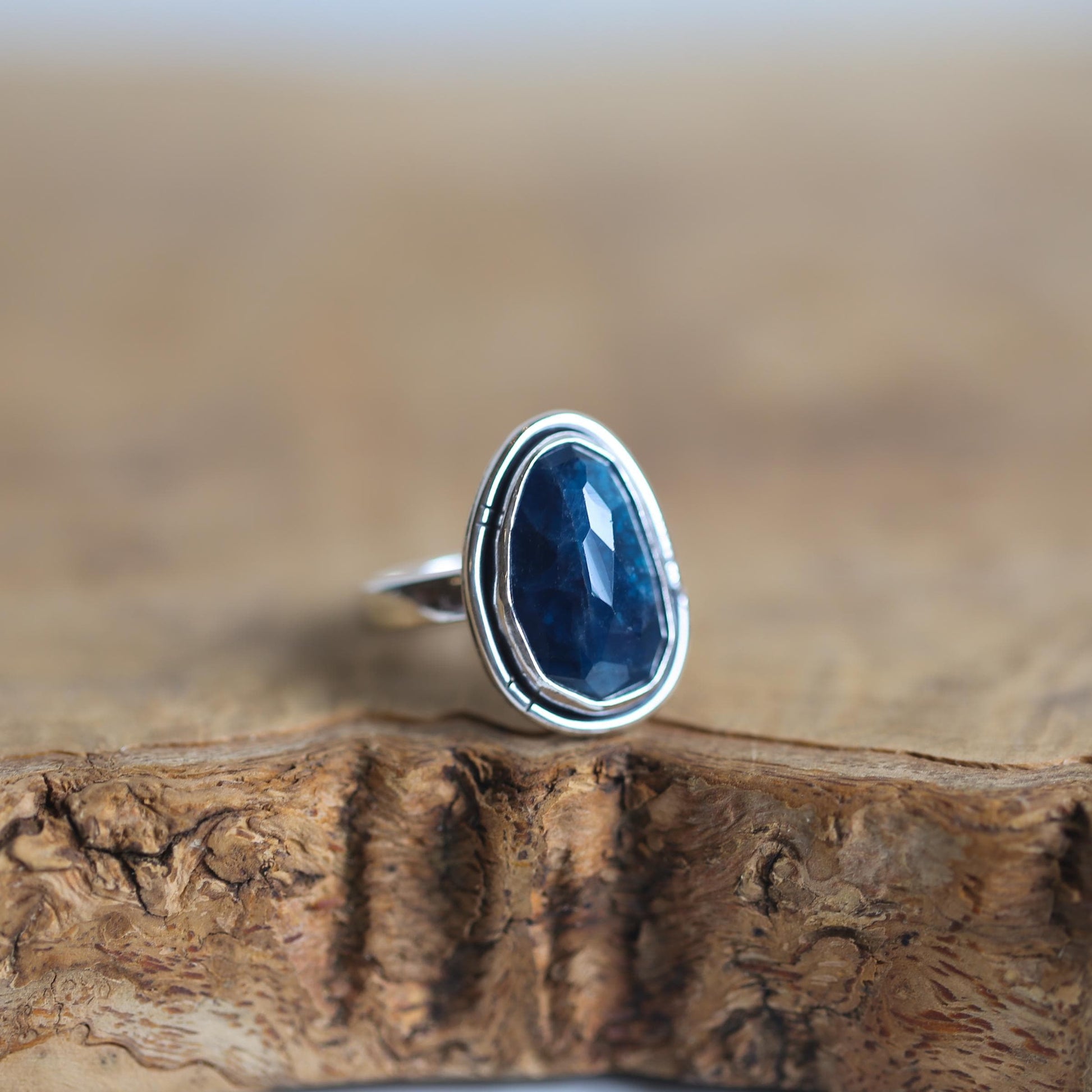 May include: A silver ring with a dark blue gemstone set in a bezel setting. The ring is on a wooden surface.