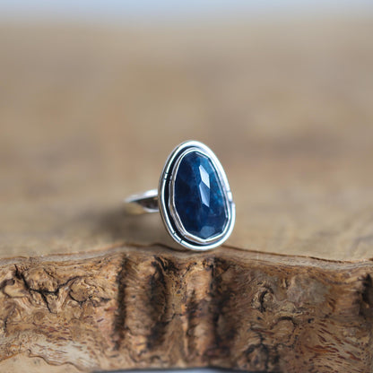May include: A silver ring with a dark blue gemstone set in a bezel setting. The ring is on a wooden surface.
