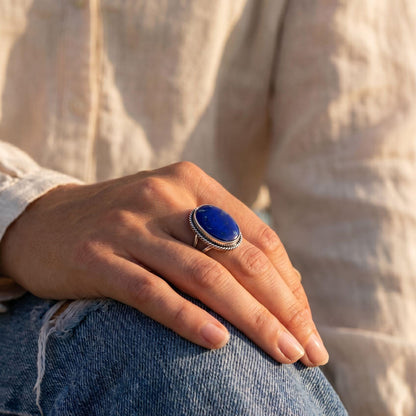 May include: A silver ring with a large, oval, deep blue gemstone. The gemstone is set in a silver band with a twisted rope design around the stone. The ring is on a person's finger, with a background of denim jeans and a light-colored shirt.