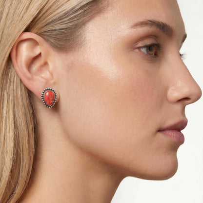 May include: A teardrop-shaped stud earring featuring a smooth, vibrant orange-red stone set in a polished silver frame. The setting is detailed with a delicate beaded border that encircles the central stone, creating a textured metallic edge.