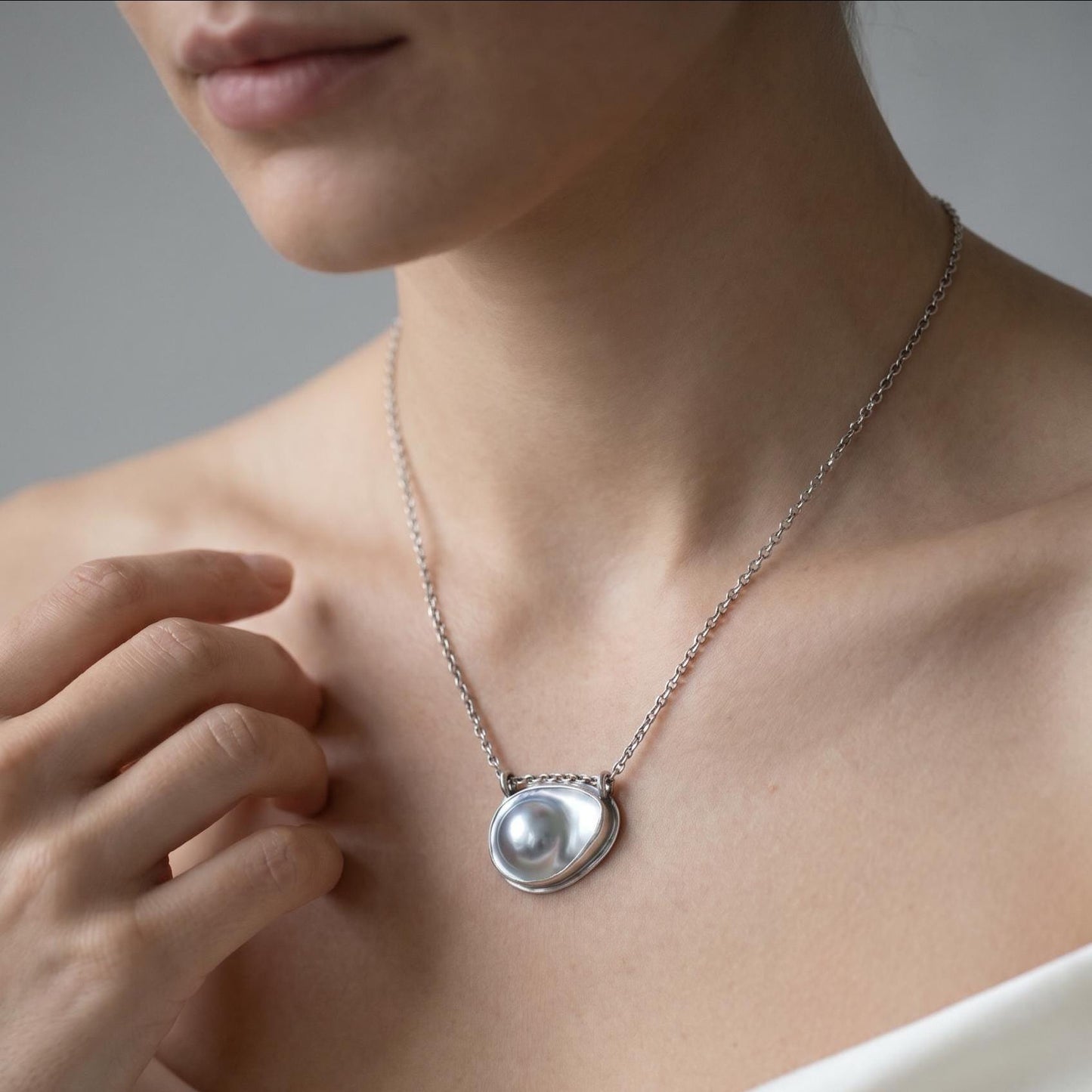 May include: A silver necklace featuring a polished, irregular, organic-shaped pendant with a central, smooth, round, lustrous pearl-like element. The pendant is suspended from a delicate, fine-linked silver chain that rests around the neck.