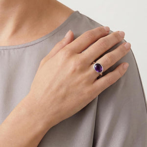 May include: A silver ring featuring a smooth, oval-shaped purple amethyst gemstone set in a polished metal band. The ring is worn on the ring finger, showcasing the deep violet color of the stone and the simple, clean design of the silver setting.