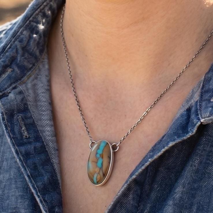 May include: A woman wearing a silver chain necklace with an oval pendant. The pendant features a turquoise and brown stone. She is wearing a denim shirt and smiling. The background shows a rodeo arena.