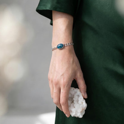 May include: A silver-toned chain link bracelet featuring a central oval-shaped blue stone setting. The bracelet is worn on a wrist, and the hand is holding a cluster of white, crystalline mineral formations with a jagged, textured surface.