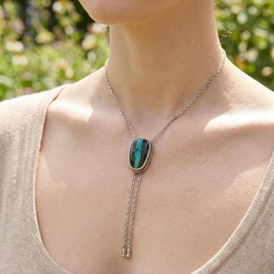May include: Silver bolo tie with an oval turquoise and black stone pendant. The pendant is framed with a silver rope design. The bolo tie has a silver chain and decorative silver tips.