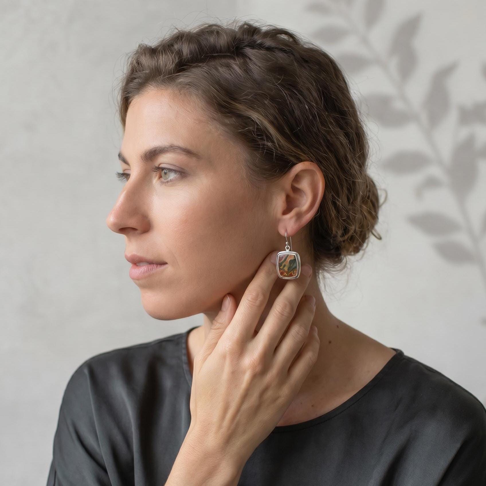 May include: A silver drop earring featuring a rectangular stone with swirling patterns of brown, green, and cream colors. The earring is suspended from a simple silver hook and has a polished metal frame surrounding the rectangular stone.