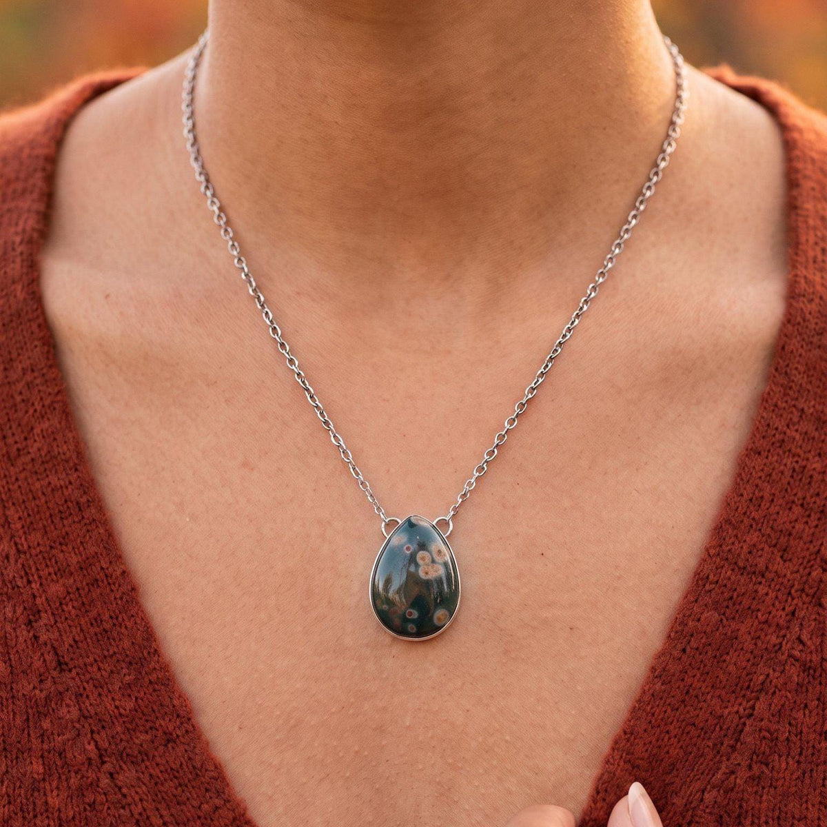 May include: A teardrop-shaped pendant necklace featuring a dark, polished stone with light-colored circular inclusions. The stone is set in a simple silver-toned metal frame and hangs from a matching silver-toned link chain around the neck.