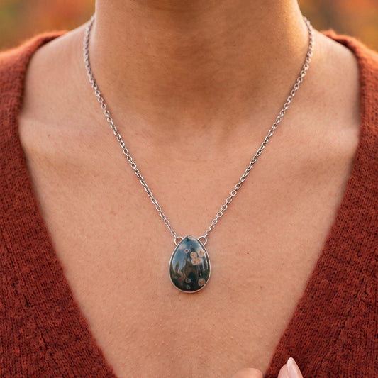 May include: A teardrop-shaped pendant necklace featuring a dark, polished stone with light-colored circular inclusions. The stone is set in a simple silver-toned metal frame and hangs from a matching silver-toned link chain around the neck.